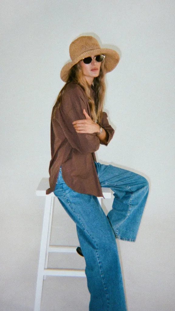 A woman sitting on a stool against a blank backdrop wearing a straw sunhat, sunglasses, brown shirt and blue jeans.